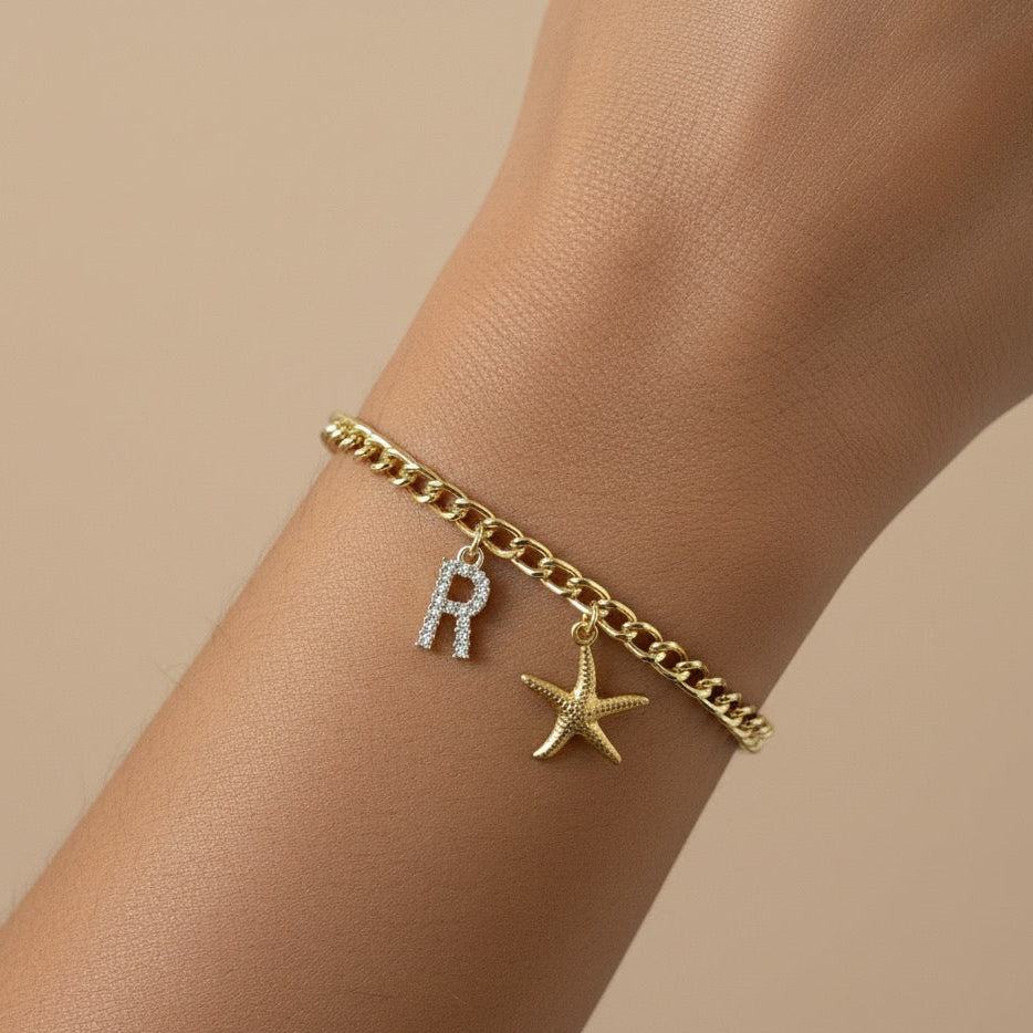 Gold bracelet with 'R' charm on a wrist against a beige background